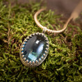 Ladda upp bild i Galleri visare, Et nærbillede af *Vedhæng - Eryn Design - Tranquill 14kt Guld* viser en stor, oval-formet grønlig-blå ædelsten sat i en indviklet sølvring. Vedhænget er lavet af Eryn Design og hænger på en flettet 14 karat guldkæde og hviler på en seng af mos. Ædelstenen reflekterer lyset og giver den et lysende udseende.