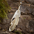 Ladda upp bild i Galleri visare, En Vedhæng Feather 925s Sølv i form af en fjer ligger på groft, barkagtigt træ med noget grønt mos synligt til venstre. Vedhænget har indviklede detaljer, med riller og snit, der efterligner den naturlige tekstur af en fuglefjer vedhæng.