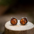 Ladda upp bild i Galleri visare, Ett par örhängen Amber Key 925s silver vilar på en träyta. De rika, varma orangebruna stenarna är infattade i en detaljerad silverram med små dekorativa pärlor, perfekt framhävd mot den suddiga bakgrunden.