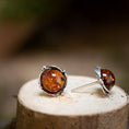 Ladda upp bild i Galleri visare, Ett par örhängen Amber Key 925s i silver med runda bärnstenar i sterlingsilver på en naturlig träyta. En står upp, den andra ligger ner och framhäver hantverket och den rödbruna tonen.