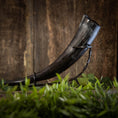 Ladda upp bild i Galleri visare, En elegant Drikkehorn Basic uden holder med en mørk nuance vises elegant på et sort metalstativ, sat mod en træbaggrund. I forgrunden introducerer sløret grønt løv et strejf af naturlig kontrast til den rustikke scene, hvilket fremkalder atmosfæren af traditionel vikingemarkeder.