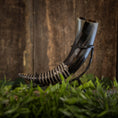 Ladda upp bild i Galleri visare, Vackert utformad Drickshorn-spiral på svart metallställ mot rustik träbakgrund. Hornet har blank, polerad yta med intrikata spiralutskärningar och omges av frodigt grönt lövverk längst ner – perfekt för vikingamarknader eller elegant dekoration.