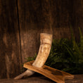 Ladda upp bild i Galleri visare, En rustik, tekstureret baggrund viser Drikkehorn Raw m. Thors Hammer Holder hvilende på et træstativ. Hornet viser en naturlig gradient af beige og brune toner, der minder om traditionel vikinge-dekoration. Grønt løv ses i baggrunden, hvilket tilføjer et jordisk præg til indstillingen.