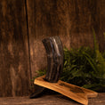 Ladda upp bild i Galleri visare, Drikkehornet Ak m. Thors Hammer Holder står oprejst på en træbase med messingdetaljer, sat mod rustikke træplanker og frodigt grønt løv, der fremkalder essensen af vikingemarkeder.