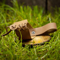 Ladda upp bild i Galleri visare, Et lille stykke solbrunt læder, der er lavet til en lille sandal, lægges på en træskive midt i levende grønt græs. Sandalen har et snøredesign, der viser detaljeret håndværk og en rustik, naturlig æstetik, perfekt til at tilføje et autentisk touch til din Drikkehorn Chief Dekoration.