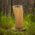 Ladda upp bild i Galleri visare, Et "Drikkeglas Horn med mønster" (300ml) med guldkant og diamantformede graveringer, fyldt med en brun væske, står på en træskranke midt i højt grønt græs med en sløret træbaggrund.