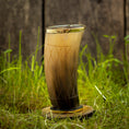 Ladda upp bild i Galleri visare, Et dekorativt Drikkeglas Horn med mønster (300 ml), med en gradient fra mørk til lysebrun og prydet med ætsede geometriske mønstre, vises på en rund træstand midt i grønt græs med et sløret træhegn i baggrunden.