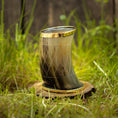 Ladda upp bild i Galleri visare, Et Drikkeglas Horn med Mønster (100ml) med indviklede udskæringer og en gylden rand er placeret oprejst på en træskive, omgivet af frodigt grønt græs. Rammerne fremstår rustikke og naturlige.