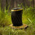 Ladda upp bild i Galleri visare, Et sort Drikkeglas Horn med Mønster (100 ml), indviklet designet med guldkant, hviler på et lille, rundt stykke træ i græsset. Baggrunden viser sløret grønt løv og en utydelig træstruktur, der fremkalder charmen og det unikke ved håndværksfremstillede stykker.