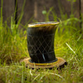 Ladda upp bild i Galleri visare, Et poleret, mørkt Drikkeglas Horn med Mønster (100ml) med en gylden kant sidder oven på en træbase på græs. Koppen har et krydsmønster ætset ind i overfladen og er sat mod en sløret baggrund med naturlige elementer.