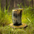 Ladda upp bild i Galleri visare, Et Drikkeglas Horn med Mønster (100ml) med en gylden kant sidder oprejst på en cirkulær træbund i græs. Hornet, der har et diamantmønster og en blank finish, er sat mod en baggrund af sløret grønt og træplanker, hvilket fremhæver dets unikke håndværk.