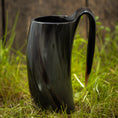 Ladda upp bild i Galleri visare, Et Vikingekrus Nature Horn (600-800 ml), med et slankt, mørkt, buet design med et håndtag, står på en plet græs. Kruset har en poleret, skinnende overflade og en organisk let snoet form. Baggrunden er lidt sløret og ser ud til at være af træ.