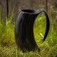 Ladda upp bild i Galleri visare, Et sort Vikingekrus Nature Horn (600-800ml) står på grønt græs med et rustikt plankeværk i baggrunden. Kruset, der efterligner den naturlige form af et dyrehorn, har en slank, blank finish og et stort buet håndtag.