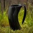 Ladda upp bild i Galleri visare, Et slankt, sort Vikingekrus Nature Horn (600-800ml) med sit unikke buede design står på græs med et plankeværk i baggrunden. Kruset har et utraditionelt håndtag, der passer problemfrit ind i dets moderne æstetik. De naturlige udendørs omgivelser står i smuk kontrast til den moderne stil på denne drikkekrus.
