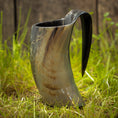 Ladda upp bild i Galleri visare, Et Vikingekrus Nature Horn (600-800ml) står oprejst på den græsklædte grund med et plankeværk i baggrunden. Hornet ligner et traditionelt vikinge-drikkekar og udviser naturlige variationer i farve, der spænder fra mørke til lysebrune nuancer.