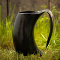 Ladda upp bild i Galleri visare, Et sort Vikingekrus Nature Horn (600-800ml) med et håndtag sidder oprejst på grønt græs, med et træhegn sløret i baggrunden. Hornet, der ligner traditionelle vikingekopper, har en blank finish og buer i en unik form.