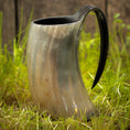 Ladda upp bild i Galleri visare, Et poleret Vikingekrus Nature Horn (600-800ml) med et håndtag står oprejst på græsklædt jord med et plankeværk i baggrunden. Hornet har en glat, reflekterende overflade med et naturligt, let gennemskinnelig mønster.
