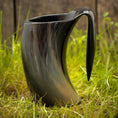 Ladda upp bild i Galleri visare, Et sort Vikingekrus Nature Horn (600-800ml) med en hornlignende form sidder på en græsklædt overflade. Denne unikke håndlavede krus har en blank finish, en bred åbning og et buet håndtag, der ligner et horn. Baggrunden er sløret, hvilket understreger kruset som fokus.