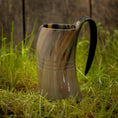 Ladda upp bild i Galleri visare, Et poleret, hornformet Vikingekrus Helleristning Horn (600-800ml) med et håndtag sidder på frodigt grønt græs. Baggrunden er en rustik trævæg, der forstærker scenens naturlige og historiske fornemmelse. Håndlavede hornkrus har subtile linjer og varierende nuancer af brun, tan og sort.