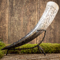 Ladda upp bild i Galleri visare, En dekorativ Drickshorn Pikto med intrikata sniderier vilar på ett svart metallstativ. Hornet har olika runsymboler och mönster etsade i ytan. Mot en rustik träbakgrund med fläckar av grön mossa skapar den vikingamarknadskänsla.