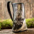 Ladda upp bild i Galleri visare, Vikingekrus Yggdrasil Horn, med et mørkt hornformet design med et hvidt keltisk knudemønster, der ligner Yggdrasil ask indgraveret på dets forside, hviler på en rustik træoverflade med en mosbeklædt baggrund, hvilket skaber en naturlig og jordnær stemning. Dette unikke krus har en rigelig kapacitet på 600-800ml.