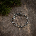 Ladda upp bild i Galleri visare, Armbånd Chainlinked 5mm 925s Sølv, lavet af oxideret 925s Sterling sølv, ligger på en forvitret træoverflade. Noget grønt mos er synligt i det øverste venstre hjørne af billedet, hvilket tilføjer et naturligt touch til den rustikke baggrund og fremhæver denne unisex-smykkes rå elegance.