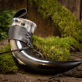 Ladda upp bild i Galleri visare, Drikkehorn Med Sort Læderholder, et poleret og buet bøffelhorn med sort og hvid marmorering, er sikkert fastgjort med en læderrem og snøre. Den hviler på en bund af grønt mos mod en rustik træbaggrund, der fremkalder vikingemarkedernes naturlige og jordnære stemning.