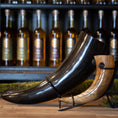 Ladda upp bild i Galleri visare, To udsøgt udformede drikkehorn, inklusive Drikkehorn Giant 1,5 liter, i forskellige størrelser og farver vises på et træbord. Hver bøffelhorn hviler på et metalstativ, og en række flasker er synlige i baggrunden på hylder. Rammerne er rustikke og elegante, der minder om traditionel vikingemarkeder.