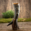 Ladda upp bild i Galleri visare, Drikkehorn Yggdrasil U. Mønster, lavet af et poleret dyrehorn, fremviser en tydelig krumning og kommer med et udsmykket udskåret stativ, der ligner det mytiske Yggdrasil-træ. Den rustikke charme ved dette stykke forstærkes yderligere af baggrunden af træplanker og grønt løv, der vækker minder om skatte fundet på et vikingemarked.