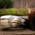 Ladda upp bild i Galleri visare, Et udskåret Drikkehorn Yggdrasil U. Mønster hviler på en træoverflade, prydet med indviklede vin- og bladgraveringer, der symboliserer Yggdrasil. Ved siden af står endnu et horn af lignende design. Baggrunden viser en rustik trætekstur med sløret grønt, der fremkalder atmosfæren af et vikingemarked.