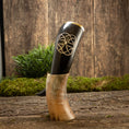 Ladda upp bild i Galleri visare, Drikkehorn Templar, et dekorativt vikinge-drikkehorn lavet af polerede bøfler med indviklede udskæringer, hviler oprejst på et træstativ. Baggrunden har en rustik træoverflade prydet med grønt mos og løv, der forstærker den naturlige og historiske æstetik, der minder om vikingemarkeder.