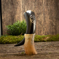 Ladda upp bild i Galleri visare, En poleret Drikkehorn Templar står oprejst i en beige meleret holder på en rustik træoverflade. Frisk grønt løv og mos fremhæver de jordiske omgivelser, og hornet har et indviklet udskåret design nær toppen, der minder om dem, man finder på vikingemarkeder.