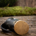 Ladda upp bild i Galleri visare, Et nærbillede af Mini Vikingekrus Horn Thors Hammer (100 ml) hviler på siden på en rustik træoverflade. Hornet, der minder om dem, der vises i Roskilde showroom, er lavet af poleret sort materiale med en lys træbase og har en let sløret naturlig grøn baggrund i baggrunden.