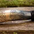 Ladda upp bild i Galleri visare, Nærbillede af Drikkehorn Yggdrasil med Mønster, der viser dens polerede overflade prydet med indviklede udskæringer. Designet har et sammenflettet knudemønster til venstre, der går over til et træmotiv med detaljerede blade i midten og til højre. Hornet hviler på en rustik træholder, der minder om vikingemarkeder.