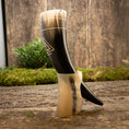 Ladda upp bild i Galleri visare, Drikkehorn Valknut, med udsøgt sort og hvid marmorering og ætset med et slående geometrisk design, hviler elegant på en naturholder træstativ. En mosbeklædt jord og træbaggrund forstærker dens naturlige, historiske charme yderligere, mens en lille bladplante tilføjer et strejf af grønt i baggrunden.