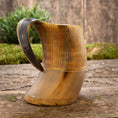 Ladda upp bild i Galleri visare, Et Vikingekrus Bark Horn (600-800ml) med træbund og håndtag hviler på en rustik træoverflade. Hornets krop er poleret med en naturlig gradient af lyse til mørkebrune nuancer, med udskårne lodrette linjer nær toppen. Grønne buske er slørede i baggrunden.