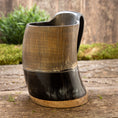 Ladda upp bild i Galleri visare, Et udsøgt håndlavet Vikingekrus Bark Horn (600-800 ml) med en trælignende tekstur på den øverste del og en glat, blank sort underdel, elegant vist på en rustik træoverflade med frodig grønt i baggrunden.
