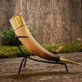Ladda upp bild i Galleri visare, Drickshorn Elegance, prytt med intrikata gulddetaljer, vilar elegant i ett slankt svart smidesjärnsstativ. Mot rustik träbakgrund och grönska framhävs hantverket och den historiska charmen, perfekt för vikingamarknader.