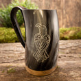Ladda upp bild i Galleri visare, Et poleret Vikingekrus Valravnen Horn (600-800 ml) har et indgraveret design af en stiliseret fugl på siden. Tankarden viser et sort håndtag og en metallisk base, der hviler elegant på en rustik træoverflade med grønt mos i baggrunden.