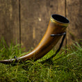 Ladda upp bild i Galleri visare, En Drikkehorn Devine, med en udsmykket messingbeklædning og en sort rem, ligger poleret på en græsklædt overflade på baggrund af et træhegn, der minder om skatte fundet på vikingemarkeder.
