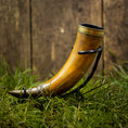 Ladda upp bild i Galleri visare, En Drikkehorn Devine med elegante metalaccenter hviler på en græsklædt overflade. Hornet er placeret oprejst og understøttet af et sort metalstativ, sat mod den rustikke kulisse af en trævæg, der minder om scener fra vikingemarkeder.