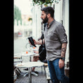 Ladda upp bild i Galleri visare, En skäggig man i grå skjorta och jeans står utanför ett café med en Gede Kaffe Drickshorn Svart. Han bär en svart axelväska och har en tatueringsärm på vänster arm. Tomma bord och stolar runt honom, med suddiga personer i bakgrunden.