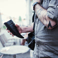 Ladda upp bild i Galleri visare, En person med tatuerade armar, i en grå skjorta, håller en Goat Kaffe Drickshorn Svart av BPA-fri plast. De står utomhus vid ett runt bord med suddig urban bakgrund. En axelrem syns på deras högra axel.
