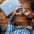 Ladda upp bild i Galleri visare, En person med tatueringar på underarmen i rutig skjorta sitter vid ett träbord. De håller en latte-kopp i ena handen och öppnar locket på sin Ged Kaffe Drickshorn Brun med den andra. Dator och kamera står också på bordet.