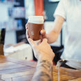 Ladda upp bild i Galleri visare, En tatuerad arm räcker ut en hand för att ta emot en get Kaffe Drickshorn Brun från en barista över cafédisken, som en viking som handlar med sitt Drickshorn. Bakgrunden är suddig och lyfter fram kaffeöverlämningen.