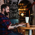 Ladda upp bild i Galleri visare, Skäggig man med tatueringar som en modern viking i röd- och blårutig skjorta skriver på bärbar dator vid ett runt träbord i ett mysigt café med bokhyllor och griffeltavlor. En getformad Kaffe Drickshorn Brun av BPA-fri plast står nära datorn.