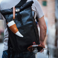 Ladda upp bild i Galleri visare, En person i grå T-shirt bär en stor svart roll-top-ryggsäck med en vit och solbrun get Kaffe Drickshorn Brun påsatt, medan han skjuter en cykel med en brun Brooks-sadel i läder. Scenen ger känslan av en modern viking på en stadsgata.