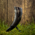 Ladda upp bild i Galleri visare, En poleret Drikkehorn Helleristning, prydet med indviklede graveringer, hviler elegant på et træstativ midt i det grønne græs og fremkalder vikingemarkedernes ånd. Et forvitret træhegn giver en rustik kulisse.