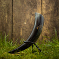 Ladda upp bild i Galleri visare, Drikkehorn Helleristning, der byder på kunstfærdige udskårne designs og en poleret finish, er elegant udstillet på et metalstativ. Den ligger på græs, med et plankeværk i baggrunden, hvilket skaber en rustik og historisk atmosfære, der minder om vikingemarkeder.