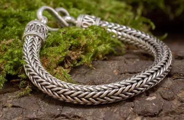 Et nærbillede af et sølvflettet armbånd med indviklede mønstre, vist på en overflade dækket af mos og bark. Armbåndet har detaljerede endestykker og en lås, der komplementerer dets vævede design.